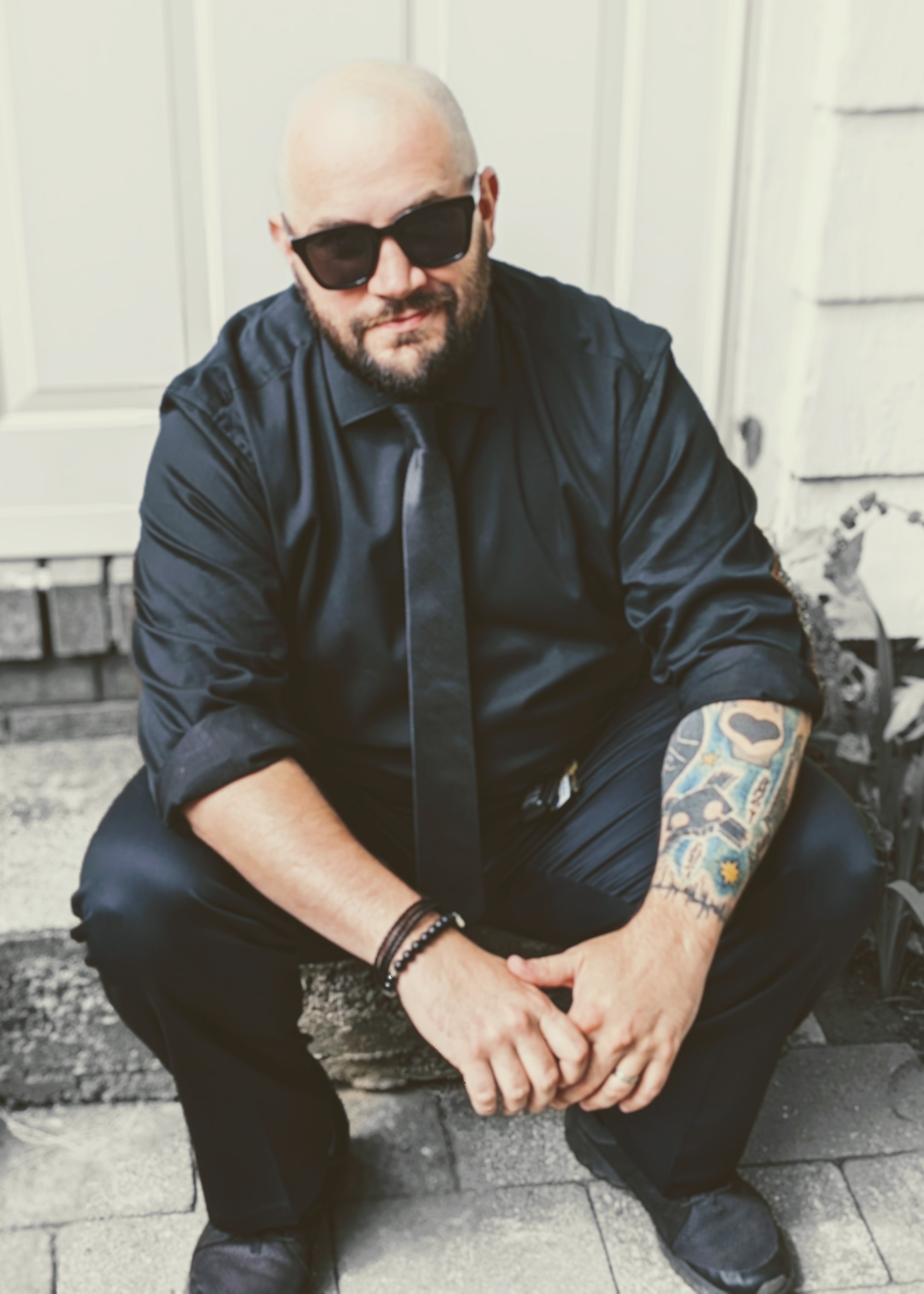 A white man sits on a front step. He is bald with a black beard and mustache. He is wearing black sunglasses, a black dress shirt and tie, black trousers, and black pants. He has a tattoo sleeve on his left arm.